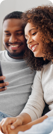 Happy couple sitting side by side, both looking at their cell phones with smiles on their faces, indicating a moment of joy.