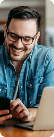 A man in a denim shirt sitting at a desk with a smartphone in his hand, in front of an open laptop.