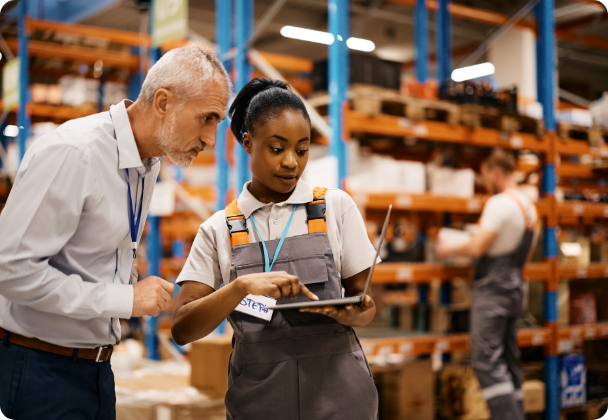 Two warehouse workers, one holding a laptop and wearing a gray apron and the other wearing a white shirt, are discussing something while standing in a warehouse with shelves stocked with goods and another worker in the background.