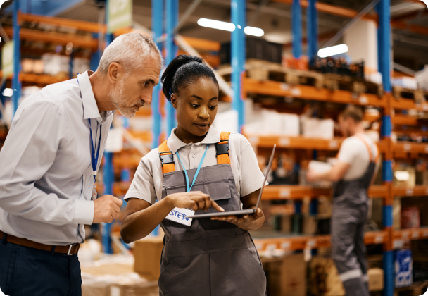 Two warehouse workers, one holding a laptop and wearing a gray apron and the other wearing a white shirt, are discussing something while standing in a warehouse with shelves stocked with goods and another worker in the background.