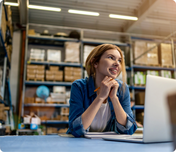 A person sitting at a desk in a warehouse with shelves of boxes in the background, in front of a laptop, appearing to be in deep thought with one hand holding a pen to a notebook.