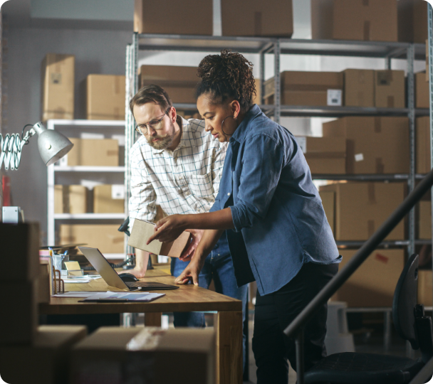 Two people working together in a warehouse, one inspecting a product with the other observing, surrounded by boxes and office equipment.